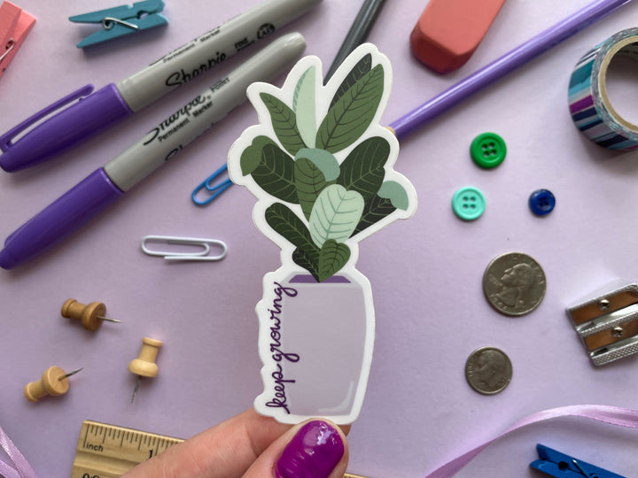 A sticker of a cartoon large-leafed green plant in a purple planter with the text "keep growing" in script up the side of the planter is held over a purple background covered in objects.