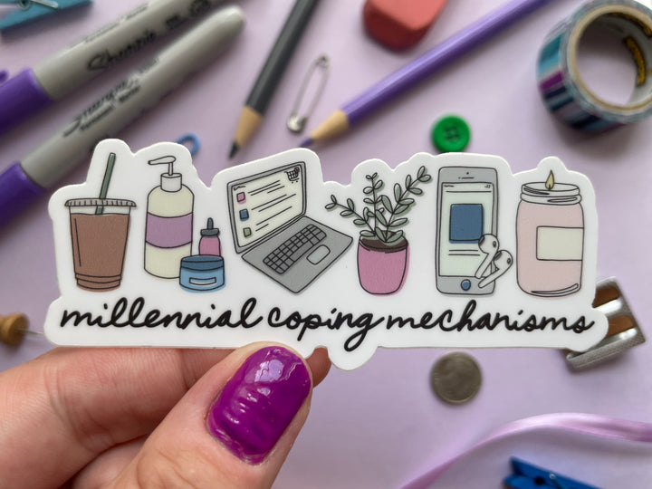 A sticker of a cartoon of a series of items including a candle, laptop, houseplant, iced coffee, lotion, and phone above the words "millennial coping mechanisms" is held over a purple background covered in objects.