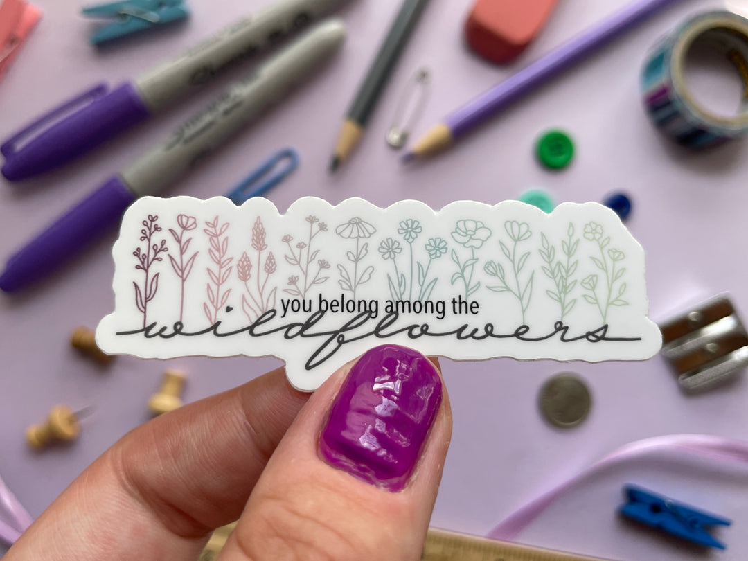 A sticker of pastel line drawings of wildflowers above the scripted text "you belong among the wildflowers" is held over a purple background covered in objects.