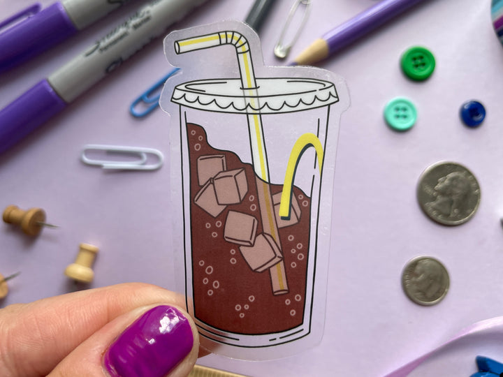 A sticker of a clear fountain drink cup with part of a yellow "M" and a bendy straw three-quarters filled with ice and bubbly brown liquid is held over a purple background covered in objects.