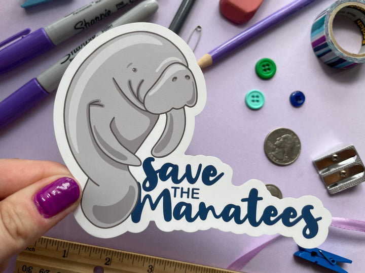 A static cling of a grey cartoon manatee standing of the size next to the dark blue text "save the manatees" is held over a purple background covered in objects.