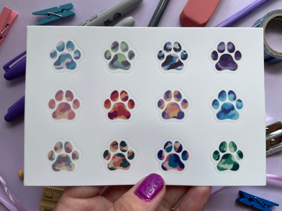 A sticker sheet of twelve multicolor paw prints is held over a purple background covered in objects.