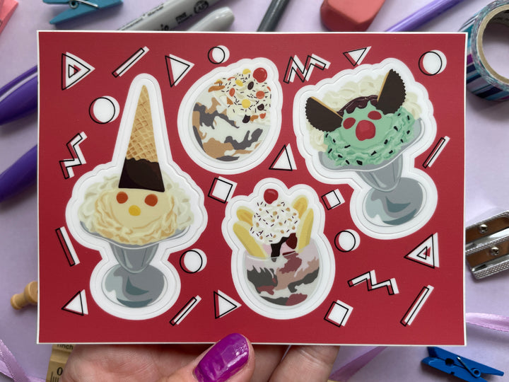 A red sticker sheet with four stickers of cartoon ice cream sundaes, two in silver parfait glasses with faces made from candy and two in glass bowls  is held over a purple background covered in objects.