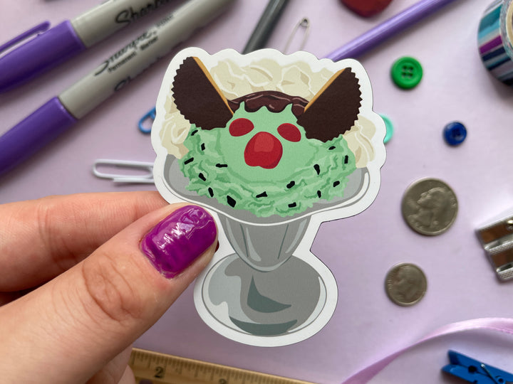 A magnet of a cartoon ice cream sundae with green ice cream and a monster face made with candy in a silver parfait glass is held over a purple background covered in objects.