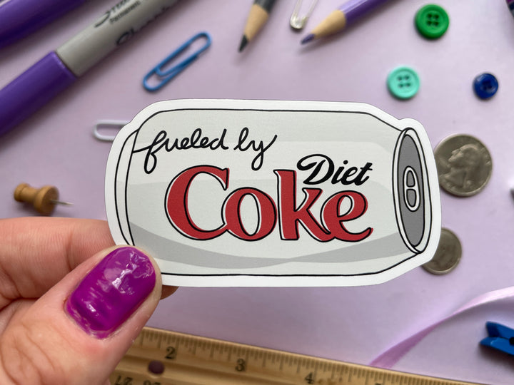 A magnet of a cartoon of a silver can of Diet Coke on its side with the text "fueled by" in the top corner is held over a purple background covered in objects.