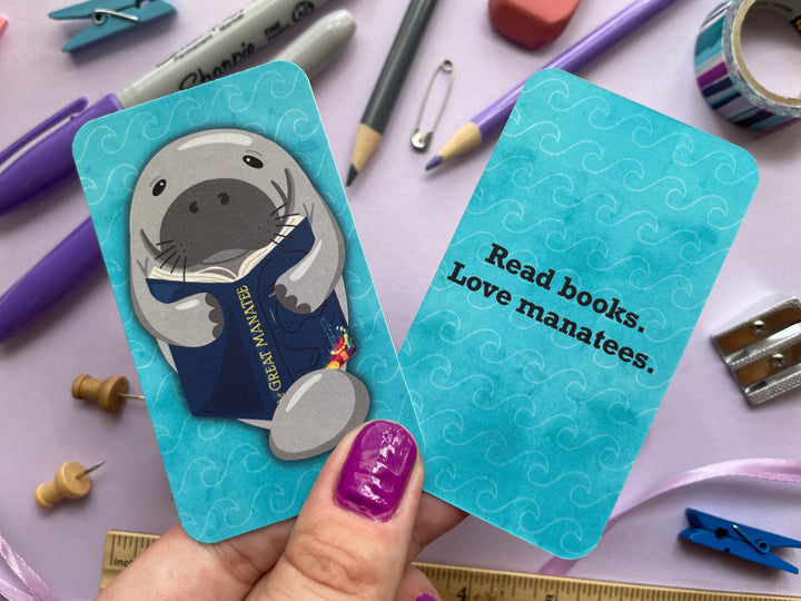 A bookmark of a grey manatee reading a book called "The Great Manatee" on the front and the text "Read Books. Love Manatees." on the back is held over a purple background covered in objects.