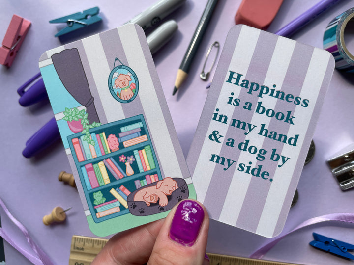 A light purple bookmark of a cartoon of an indoor scene with a bookshelf and sleeping puppy on the front and the text "happiness is a book in my hand & a dog by my side" on the back is held over a purple background covered in objects.