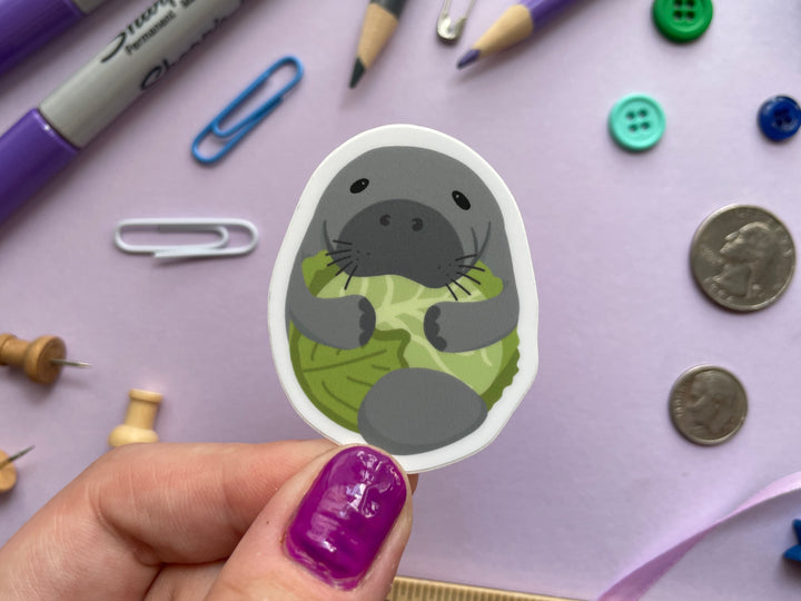 A sticker of a cartoon grey manatee with flippers wrapped around a head of green lettuce is held over a purple background covered in objects.