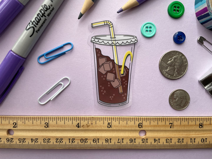 A sticker of a clear fountain drink cup with part of a yellow "M" and a bendy straw three-quarters filled with ice and bubbly brown liquid is shown on a purple background surrounded by small objects.