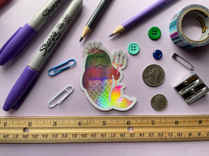 A holographic sticker of a cartoon grey manatee with a multi colored mermaid tail and yellow crown holding a yellow trident is shown on a purple background surrounded by small objects.