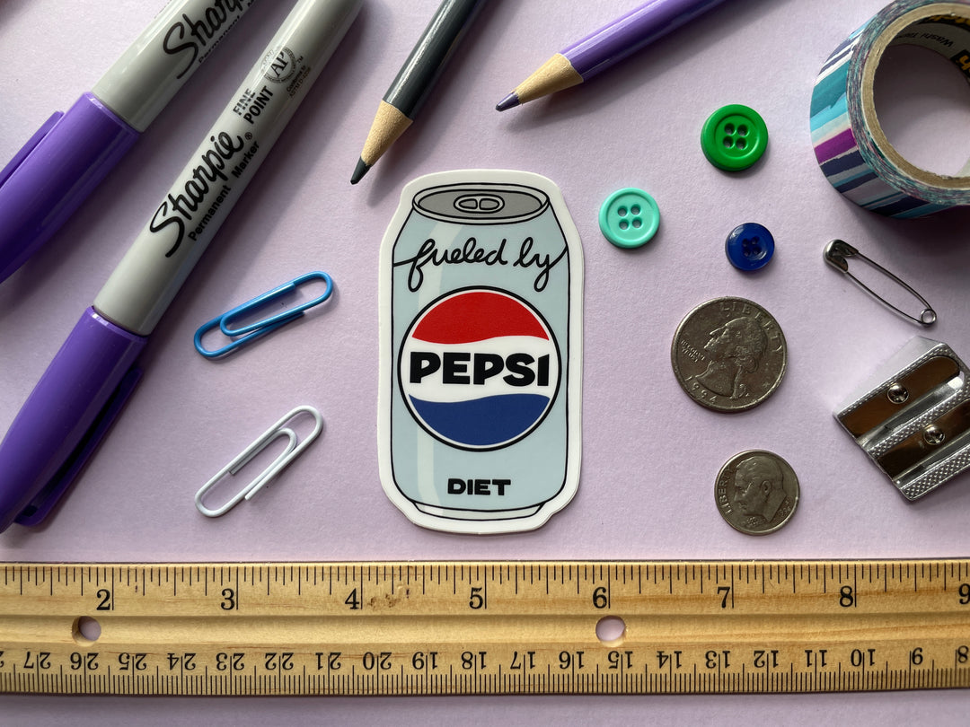 A sticker of a cartoon of a light blue can of Diet Pepsi with the text "fueled by" in the top corner is shown on a purple background surrounded by small objects.