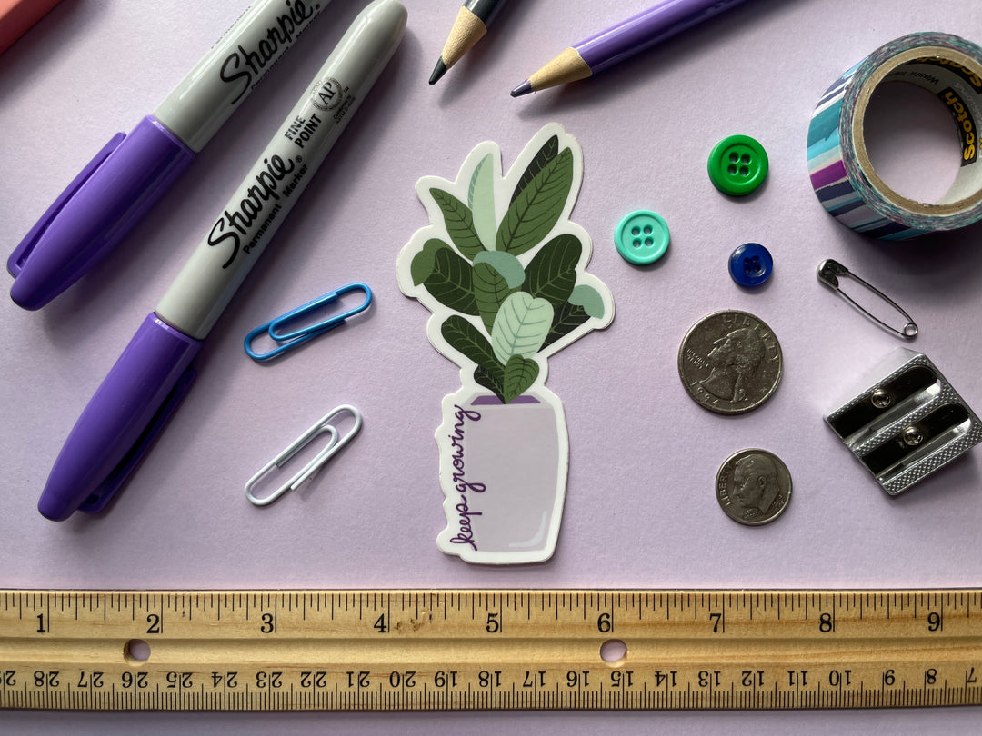 A sticker of a cartoon large-leafed green plant in a purple planter with the text "keep growing" in script up the side of the planter is shown on a purple background surrounded by small objects.