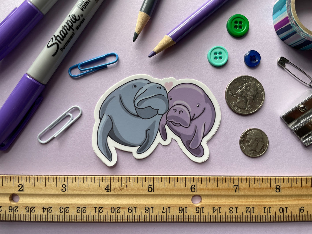 A sticker of a cartoon pair of manatees, one blue and one purple, with their heads together and a heart about them is shown on a purple background surrounded by small objects.