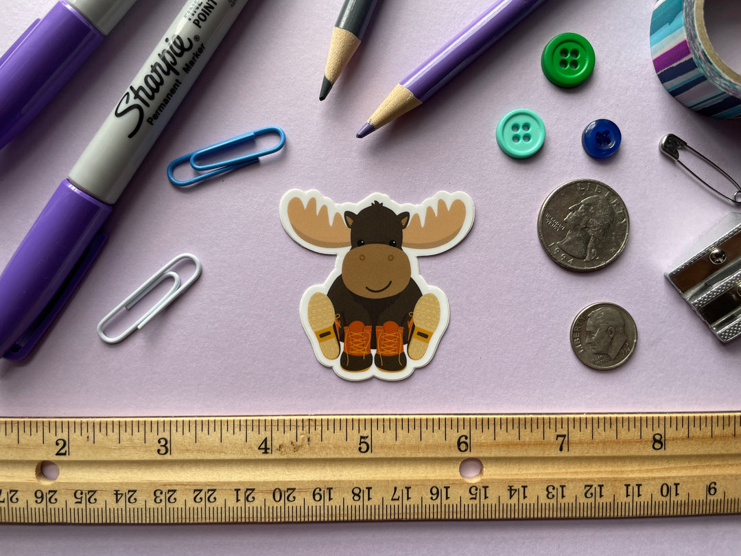 A sticker of a smiling cartoon moose sitting with orange and brown boots on its hooves is shown on a purple background surrounded by small objects.