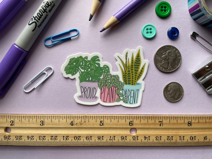 A sticker of a cartoon of three house plants in purple, pink, and blue planters with the text "proud plant parent" is shown on a purple background surrounded by small objects.