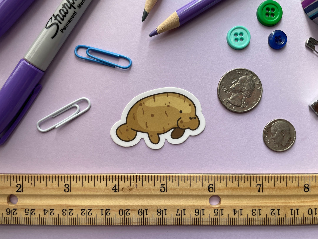 A sticker of a manatee shown in profile colored like a potato is shown on a purple background surrounded by small objects.