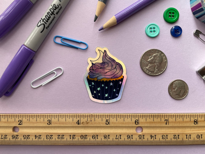 A holographic sticker of a cartoon of a cupcake with starry paper, purple and blue stardust frosting, and top with a crescent moon is shown on a purple background surrounded by small objects.