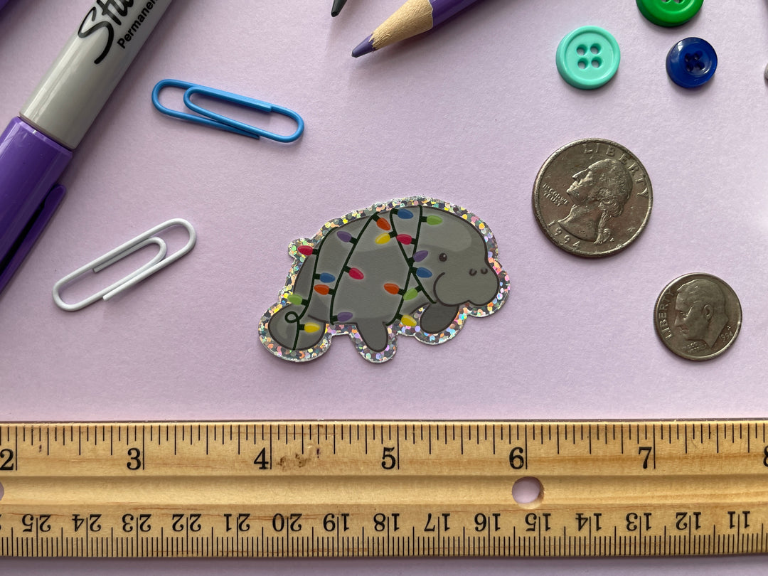 A glittery sticker of a cartoon grey manatee shown in profile wrapped in multi-color holiday lights is shown on a purple background surrounded by small objects.