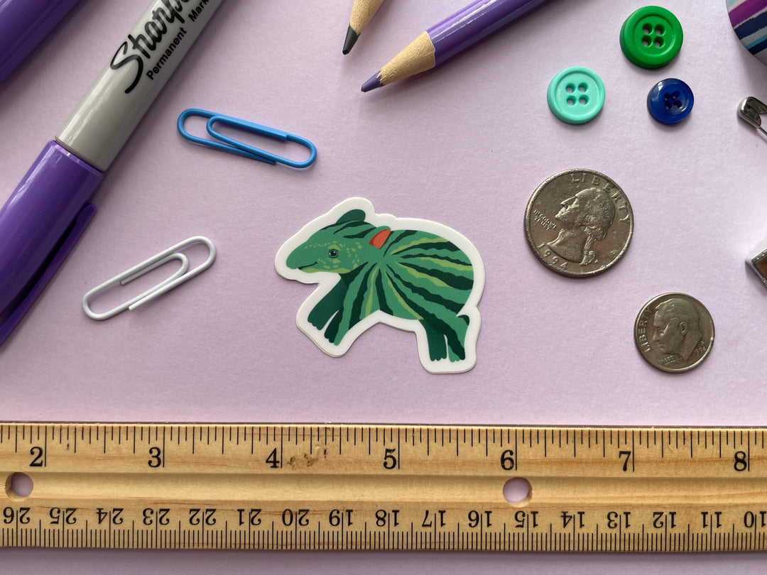 A sticker of a green cartoon tapir with watermelon stripes along its body is shown on a purple background surrounded by small objects.