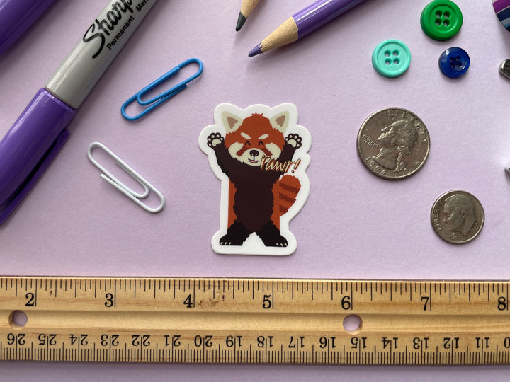 A sticker of a cartoon red panda standing on its hind legs with its arms raised and the text "rawr" next to its mouthis shown on a purple background surrounded by small objects.