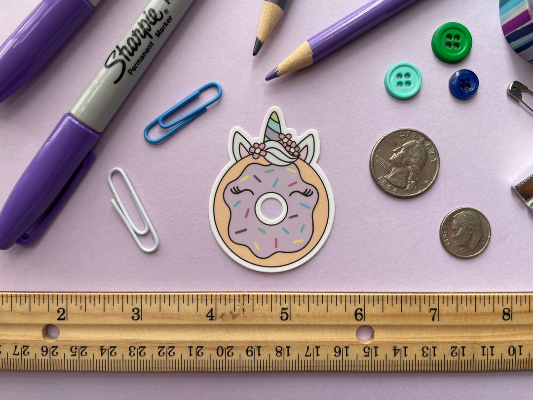 A sticker of a cartoon donut with pink frosting with horse ears and a green and blue striped unicorn horn is shown on a purple background surrounded by small objects.