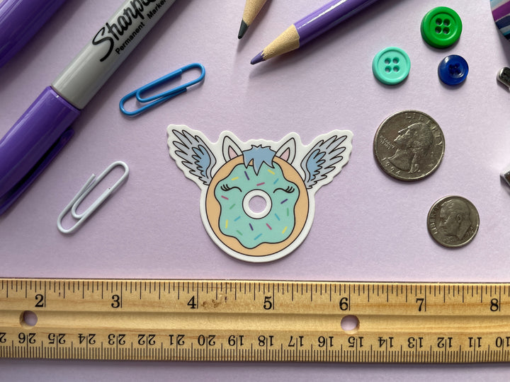 A sticker of a cartoon donut with blue frosting with blue feathered wings and horse ears and mane is shown on a purple background surrounded by small objects.