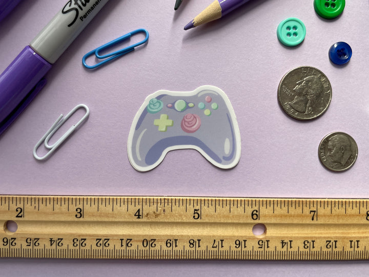 A sticker of a cartoon purple game controller with multi color buttons is shown on a purple background surrounded by small objects.