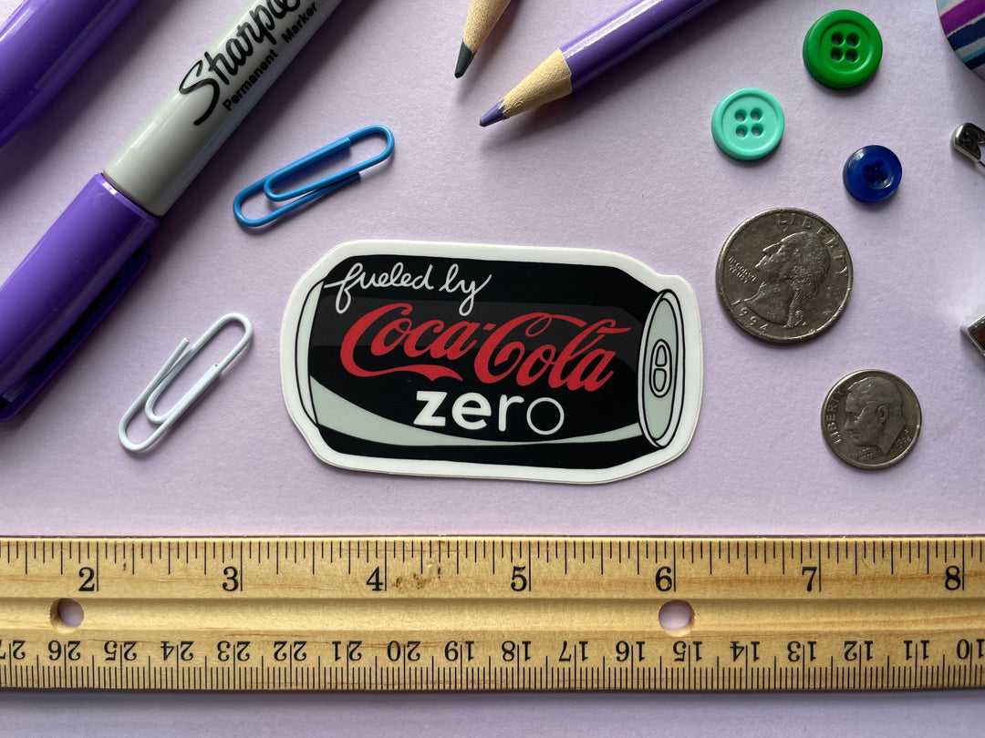 A sticker of a cartoon of a black can of Coke Zero on its side with the text "fueled by" in the top corner is shown on a purple background surrounded by small objects.