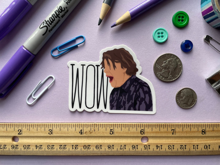 A sticker of a cartoon of comedia Alan Davies with his mouth opening shouting "Wow" from the TV show Taskmaster is shown on a purple background surrounded by small objects.