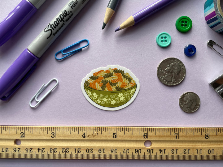 A sticker of a green bowl of orange, green, and cream colored spiraled rotini noodles is shown on a purple background surrounded by small objects.