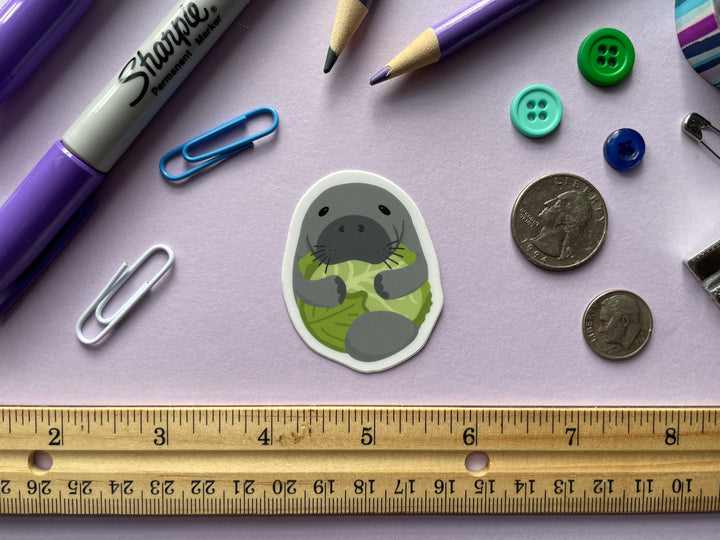 A sticker of a cartoon grey manatee with flippers wrapped around a head of green lettuce is shown on a purple background surrounded by small objects.