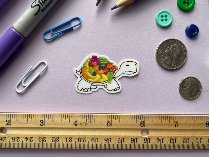 A sticker of a cartoon turtle drawn in black and white with a colorful shell of various fruits is shown on a purple background surrounded by small objects.