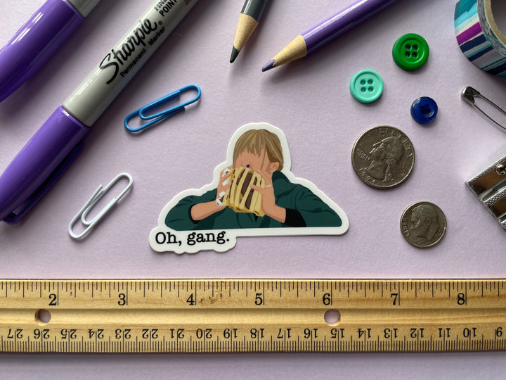 A sticker of a cartoon of comedian Mel Giedroyc eating a sandwich of candy with a blue candy in her nose from the TV show Taskmaster is shown on a purple background surrounded by small objects.