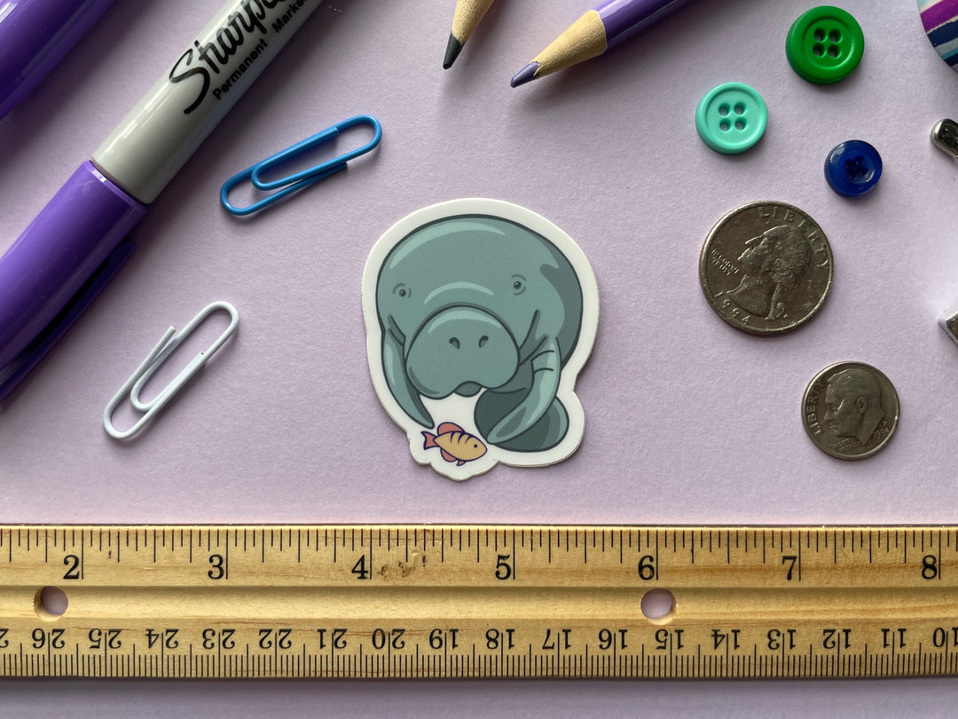 A sticker of a cartoon blue manatee head on with a yellow and orange fish beneath it is shown on a purple background surrounded by small objects.