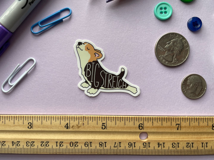 A sticker of a cartoon corgi stretching its back legs behind it with the text "big stretch" on top is shown on a purple background surrounded by small objects.