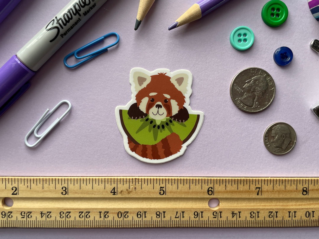 A sticker of a cartoon red panda holding a large slice of kiwi fruit is shown on a purple background surrounded by small objects.