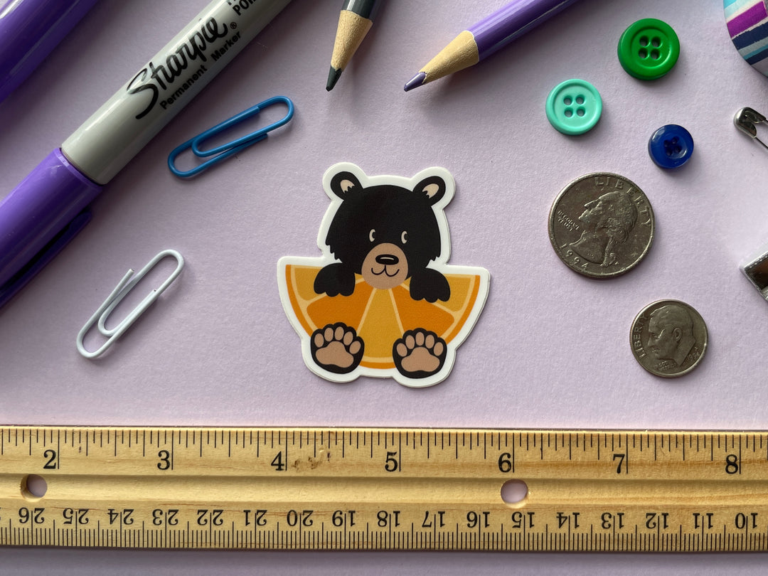 A sticker of a cartoon black bear holding a large orange fruit slice is shown on a purple background surrounded by small objects.