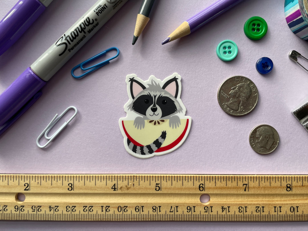 A sticker of a cartoon raccoon holding a large slice of red apple is shown on a purple background surrounded by small objects.