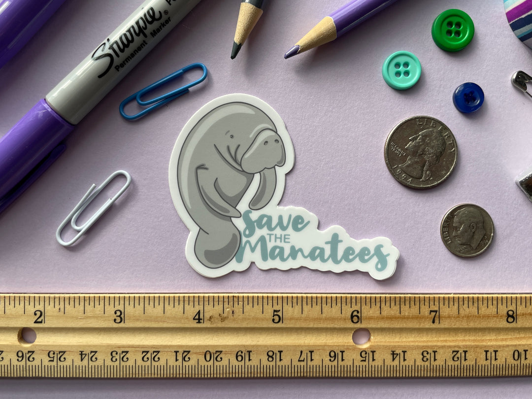 A sticker of a grey cartoon manatee standing of the size next to the light blue text "save the manatees" is shown on a purple background surrounded by small objects.