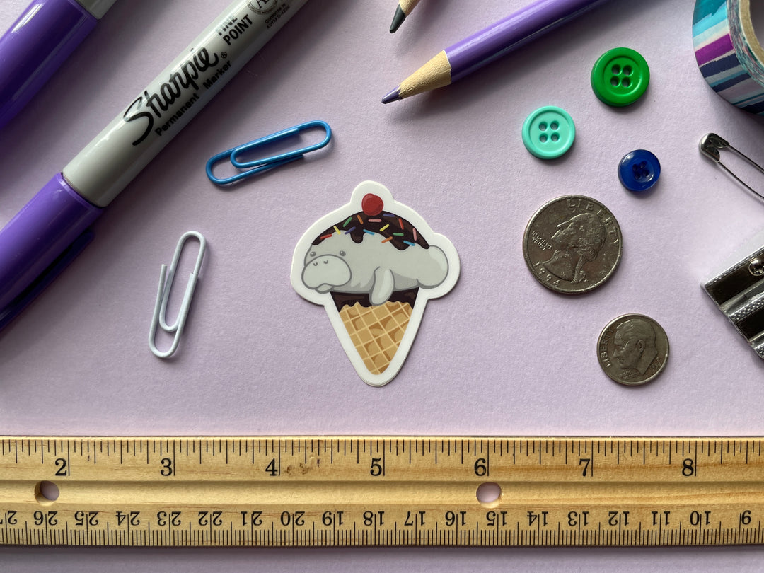 A sticker of a cartoon ice cream cone dipped in chocolate topped with a grey manatee with chocolate sauce, sprinkles, and a cherry is shown on a purple background surrounded by small objects.