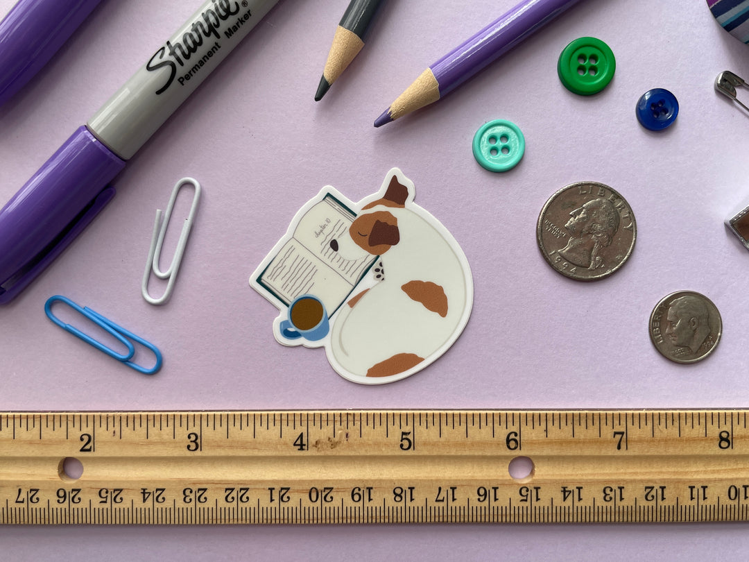 A sticker of a brown and white dog curled up next to an open box and mug of brown liquid is shown on a purple background surrounded by small objects.