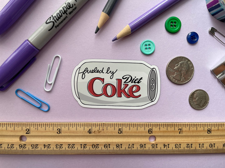 A magnet of a cartoon of a silver can of Diet Coke on its side with the text "fueled by" in the top corner is shown on a purple background surrounded by small objects.