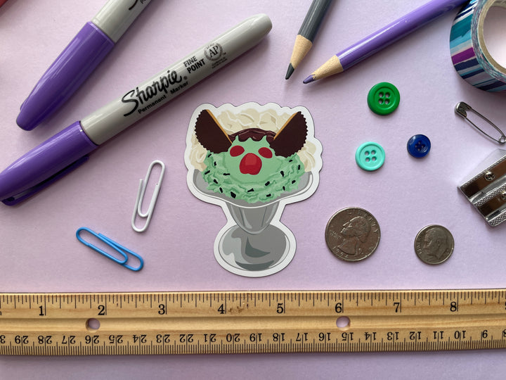 A magnet of a cartoon ice cream sundae with green ice cream and a monster face made with candy in a silver parfait glass is shown on a purple background surrounded by small objects.