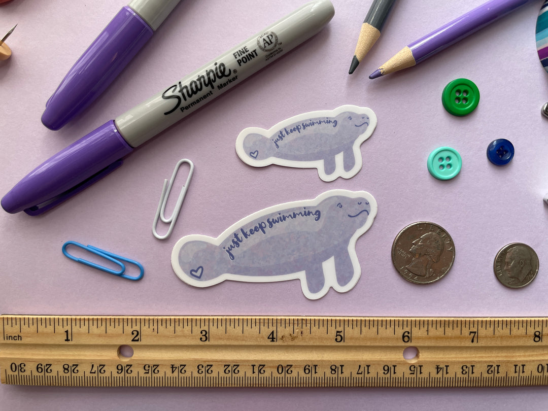 A sticker of a cartoon light purple smiling manatee shown from the side with the text "just keep swimming" over top is shown on a purple background surrounded by small objects.