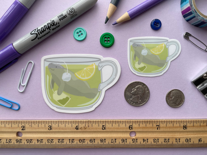 Two stickers, both of a cup of green tea with a tea bag and slice of lemon with a grey manatee swimming in it, are shown on a purple background surrounded by small objects.