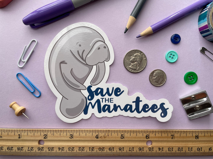 A static cling of a grey cartoon manatee standing of the size next to the dark blue text "save the manatees" is shown on a purple background surrounded by small objects.