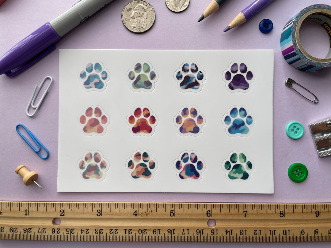 A sticker sheet of twelve multicolor paw prints is shown on a purple background surrounded by small objects.