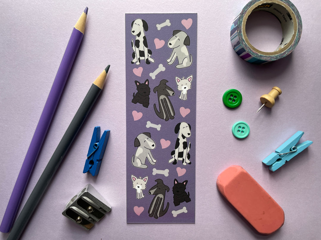 A purple bookmark covered in pink hearts and black and white drawings of cartoon dogs is shown on a purple background surrounded by small objects.