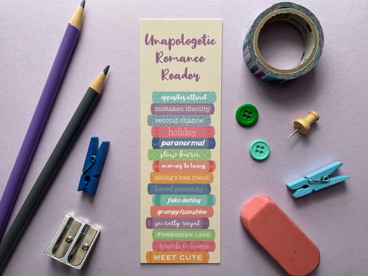 A tan bookmark of a stack of multicolored books with different romance tropes along the spines and the text "Unapologetic Romance Reader" along the top is shown on a purple background surrounded by small objects.