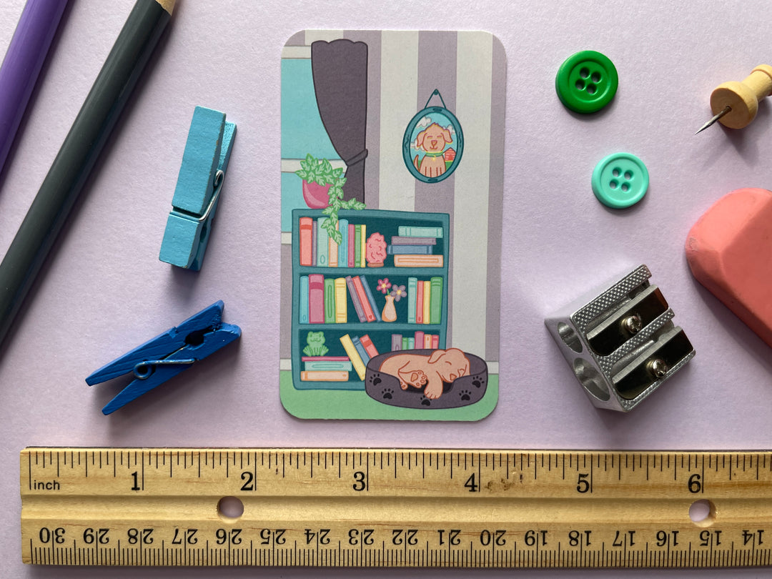 A light purple bookmark of a cartoon of an indoor scene with a bookshelf and sleeping puppy on the front and the text "happiness is a book in my hand & a dog by my side" on the back is shown on a purple background surrounded by small objects.